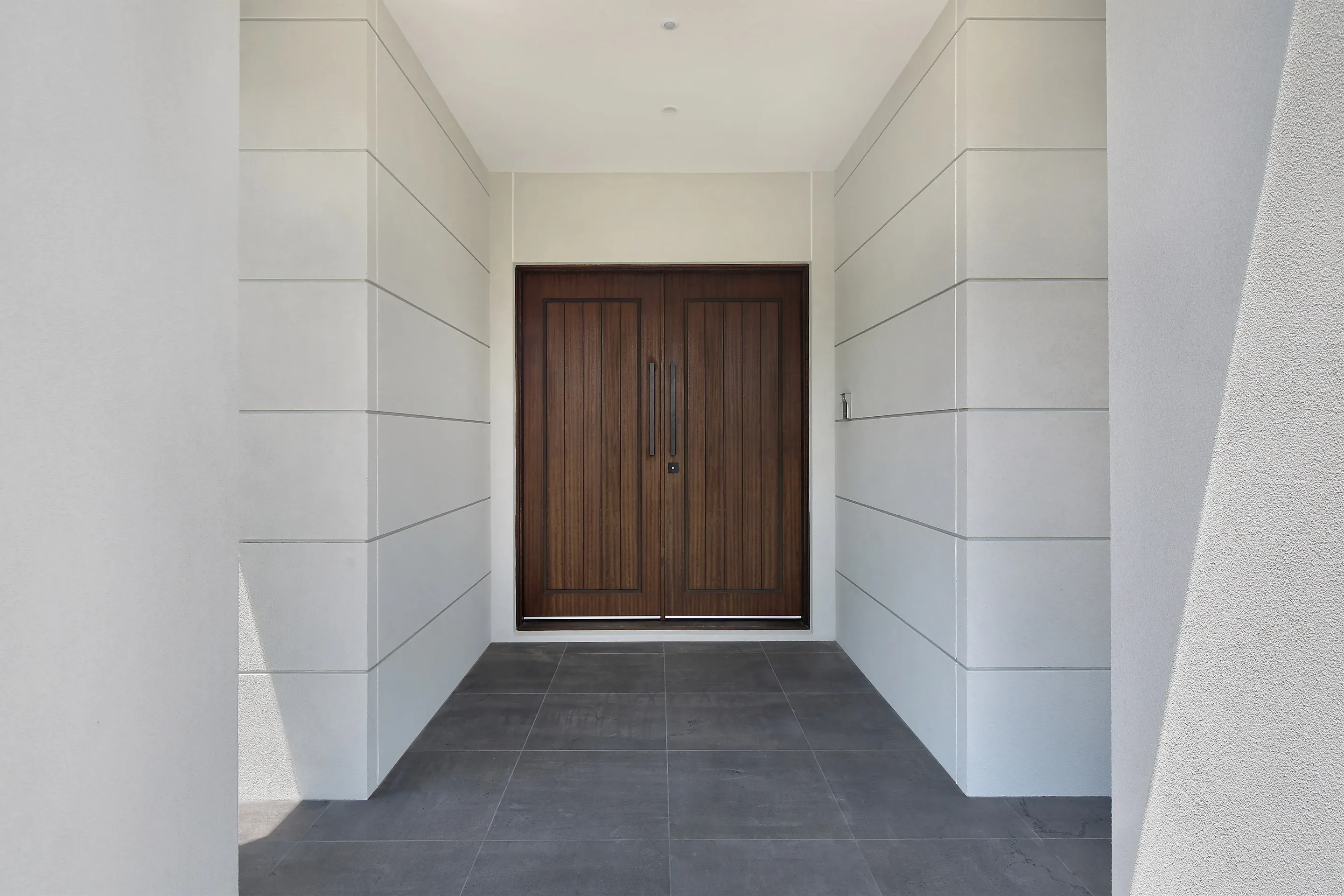 Entry portico of Essendon Residence with horizontally scored rendered walls, dark-stained timber double doors and a dark stone-tiled floor