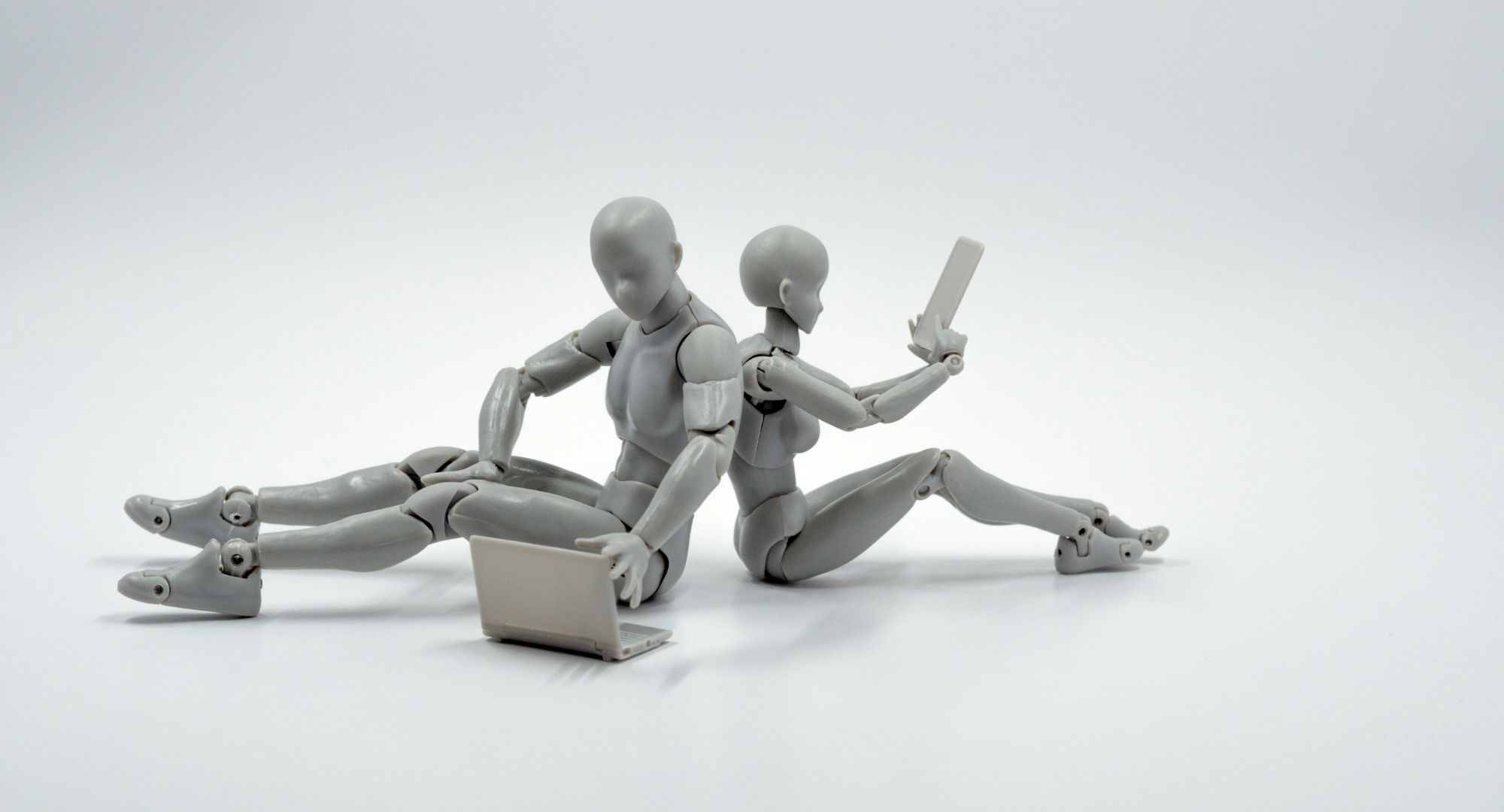 dois robôs cinzas sentados de costas, o da esquerda está digitando em um notebook enquanto o outro em um celular, fundo branco