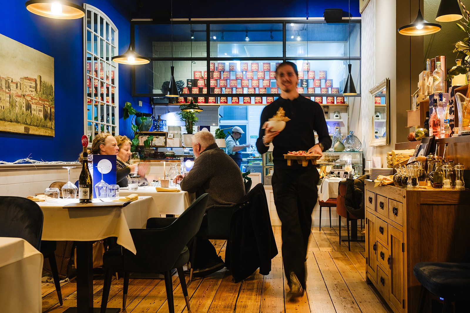 A lively restaurant scene with a seated diner in the foreground and a server walking past holding food, slightly blurred in motion. Behind them, a chef works in an open kitchen framed by a wall of stacked red and yellow “POMO D’ORO” boxes, with warm lighting and plants adding to the cosy atmosphere.