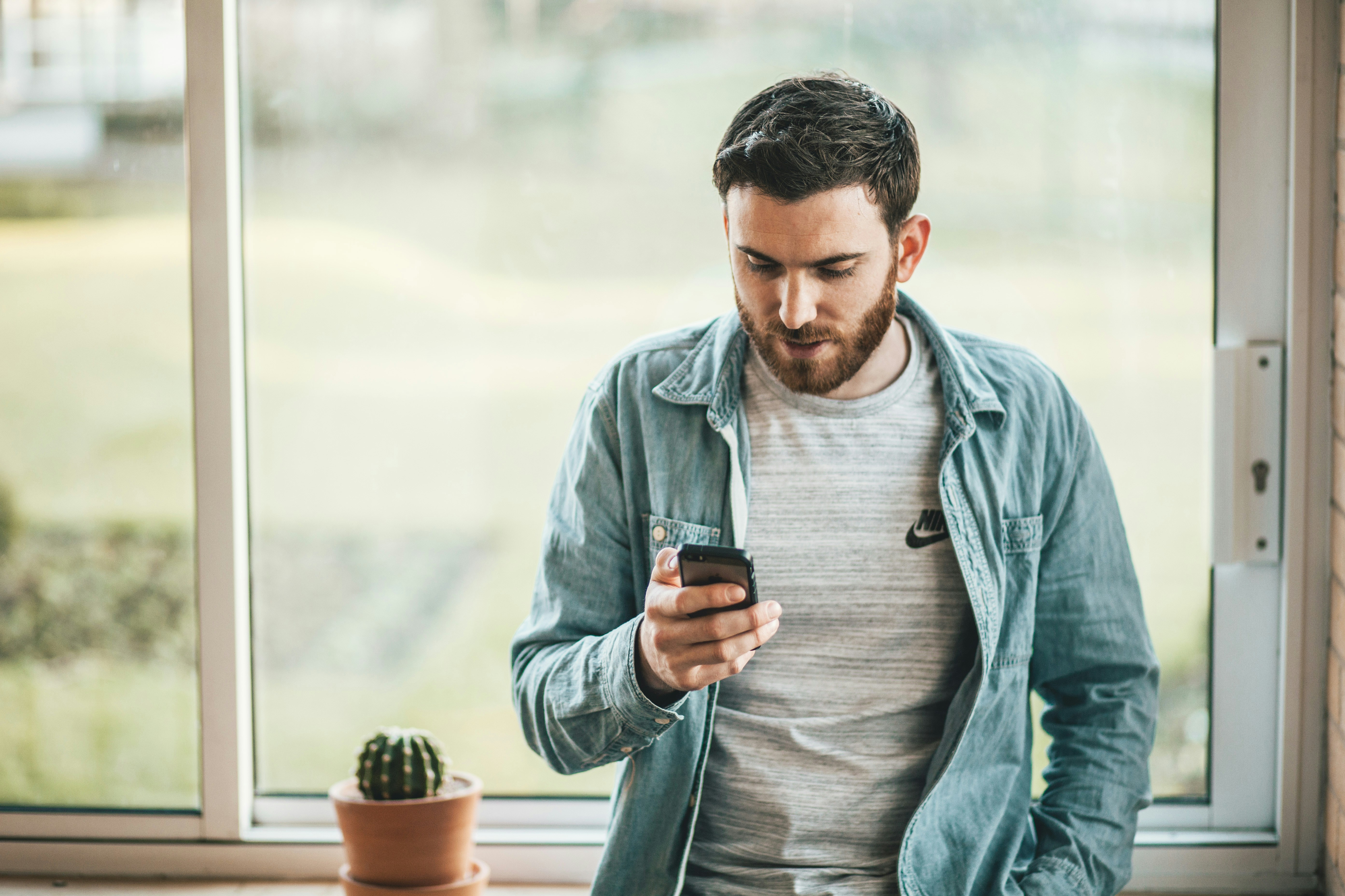 A man leans against a window and looks down at his phone.