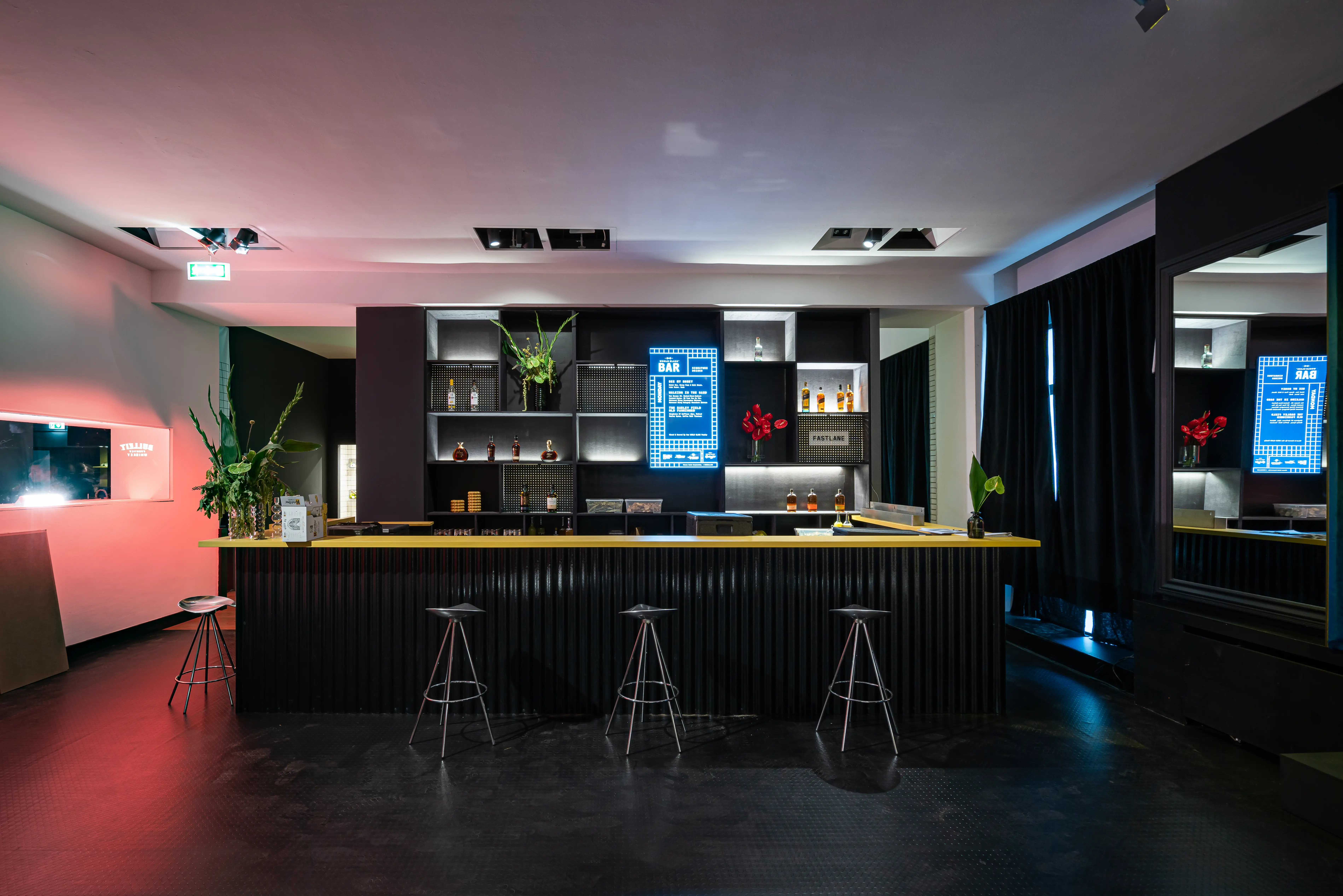 Dark bar at DIAGEO BCB19 with ribbed black counter, yellow top edge, bar stools and backlit shelves with blue menu screens under pink light.