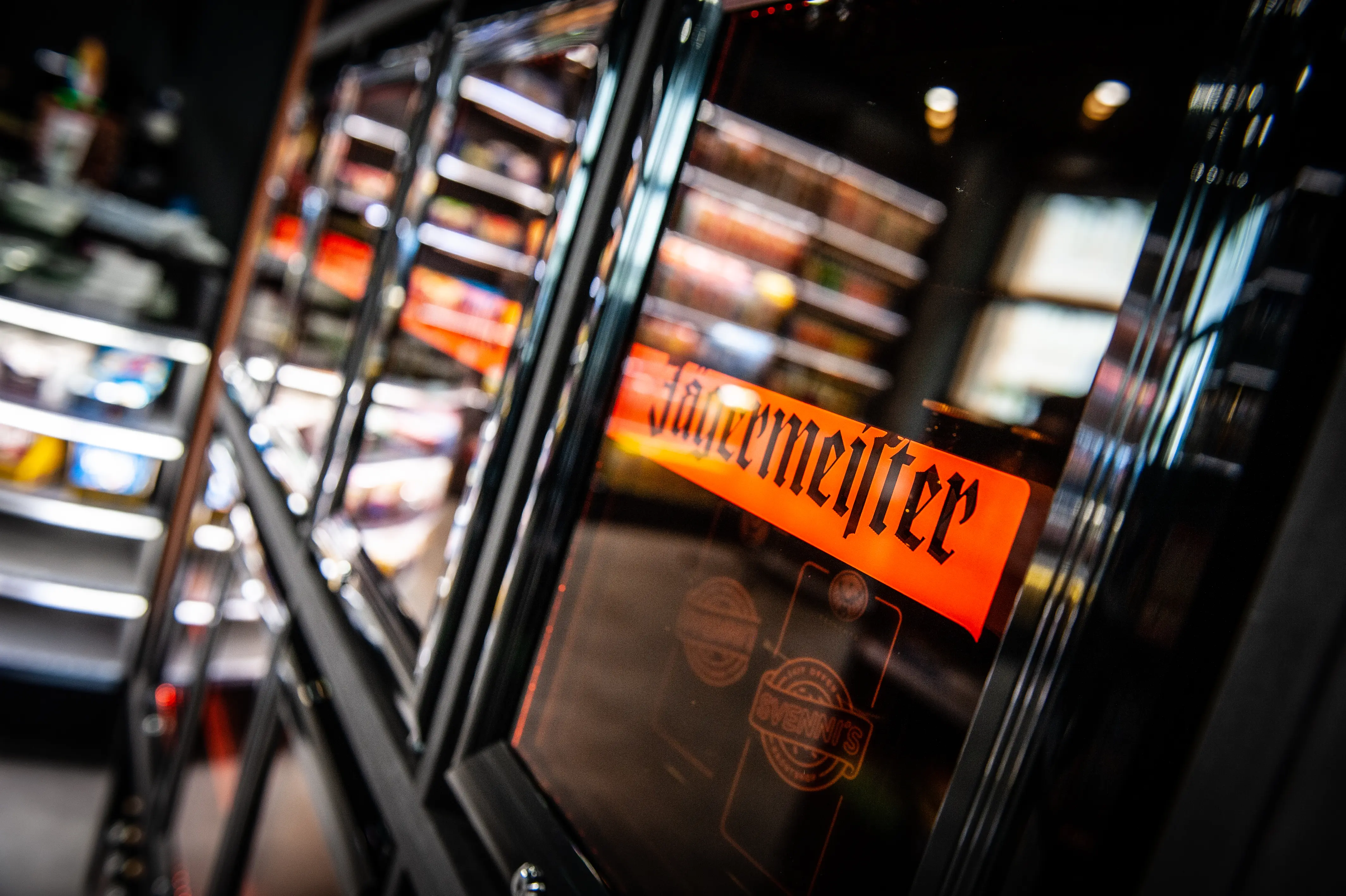Close-up of a glass fridge door with orange Jägermeister lettering at JÄGERMEISTER x SVENNI'S, with blurred kiosk shelves in the background.