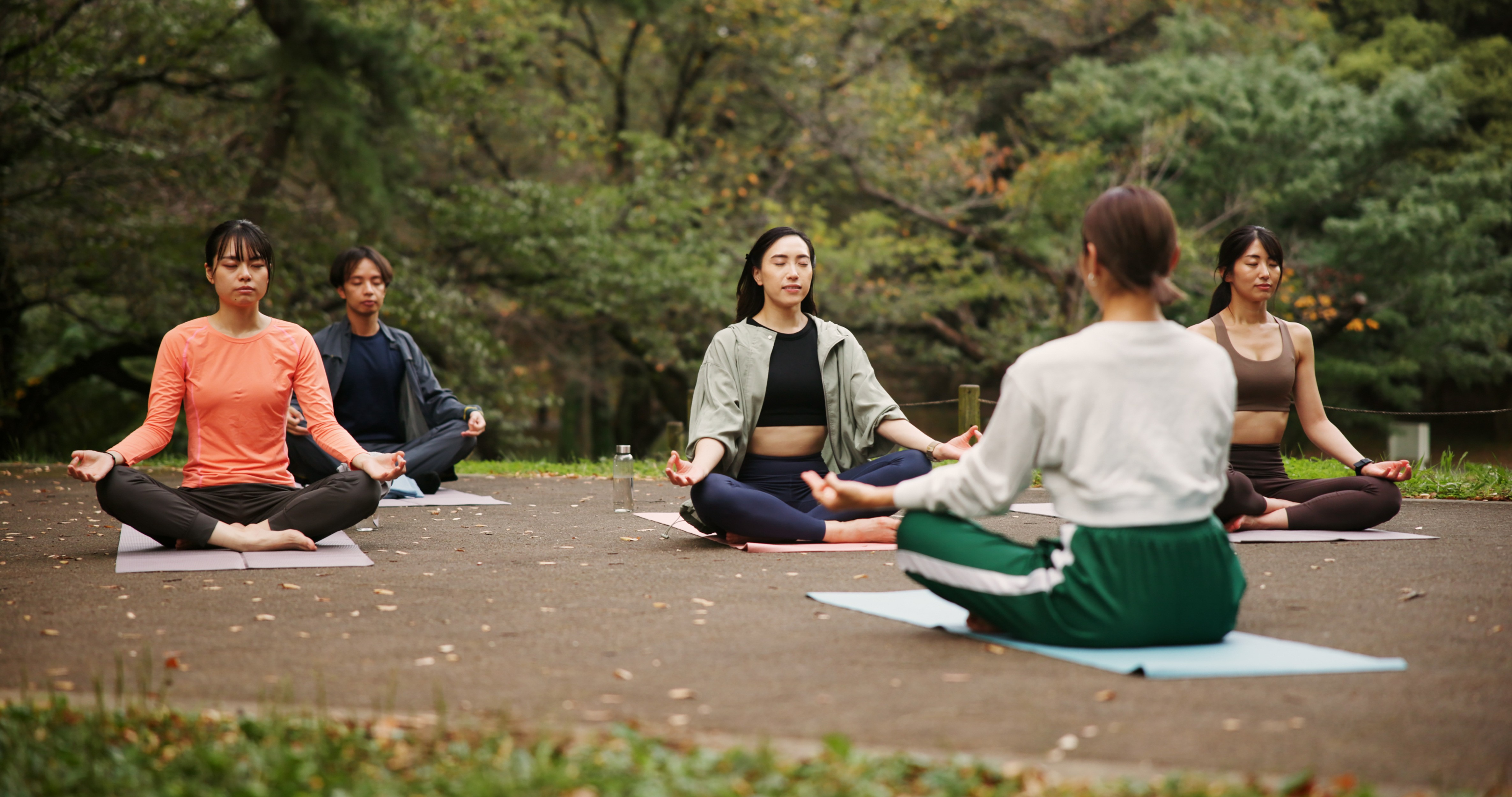 A group of people sitting cross-legged on yoga mats in a circle, practicing outdoor meditation led by an instructor in a park surrounded by trees.
