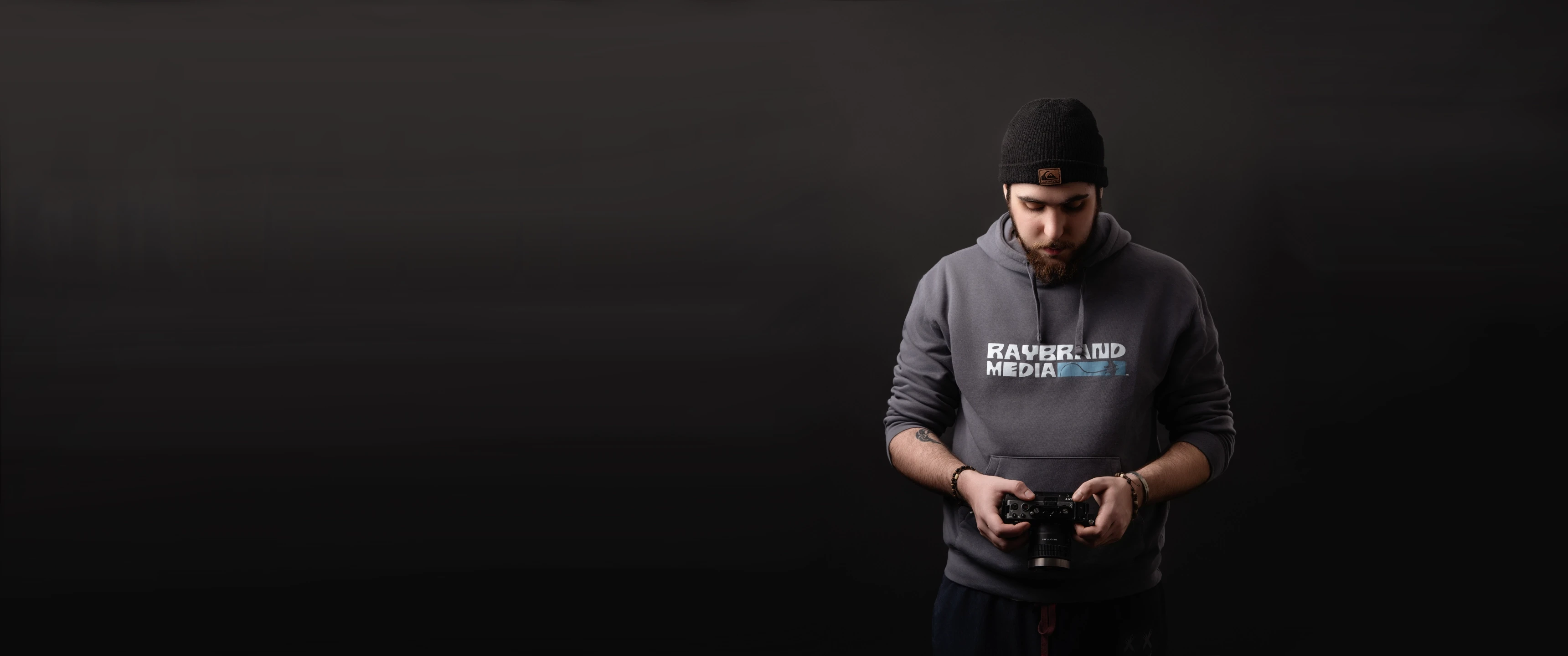 Videographer wearing Raybrand Media hoodie adjusting a camera against a dark background, representing professional video production, content creation and personal brand identity