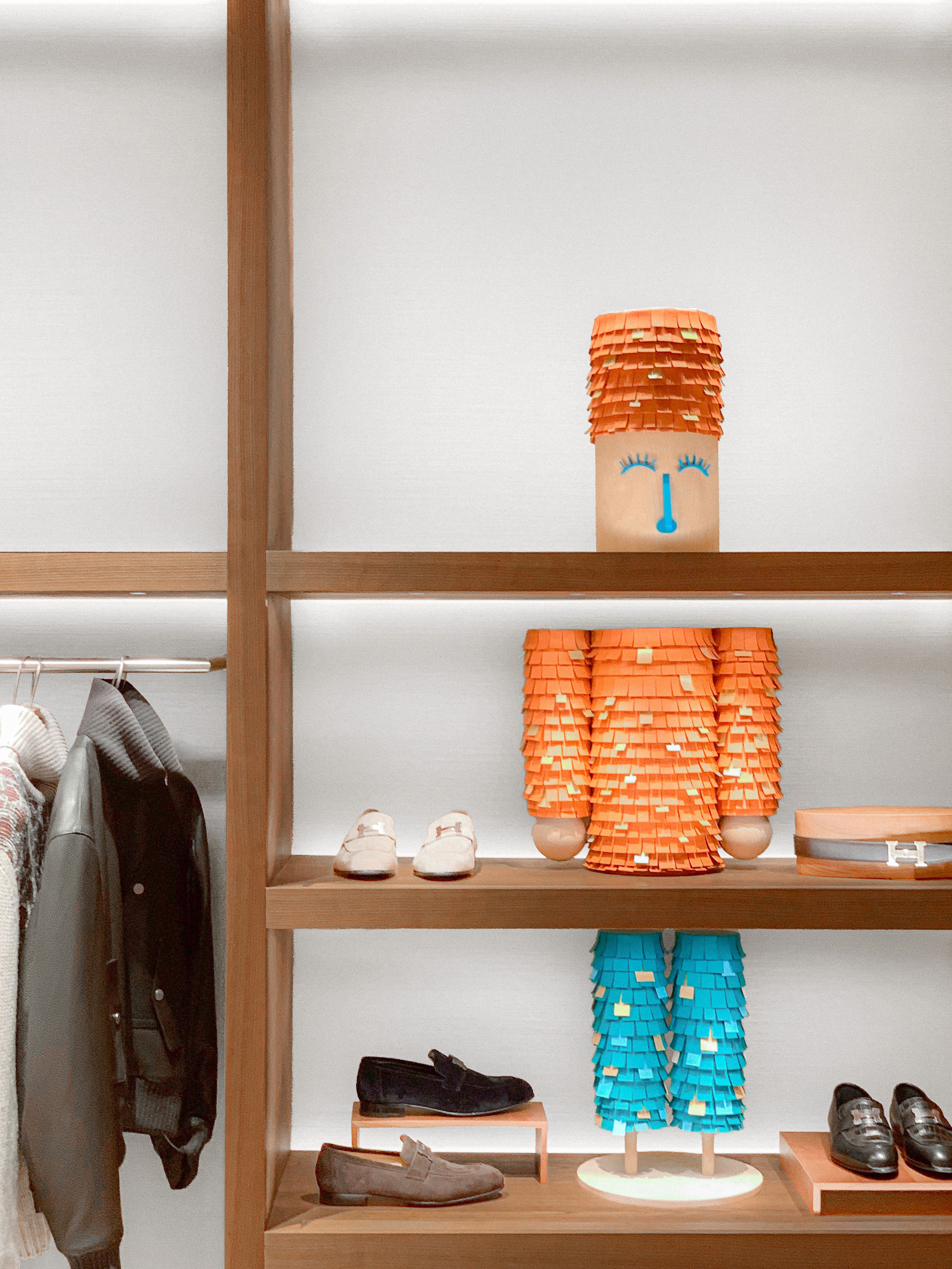 In-store display for the Hermès holiday scenography featuring wooden shelves with menswear and a playful character made from orange, yellow, and blue fringed paper, designed by WM Paris.