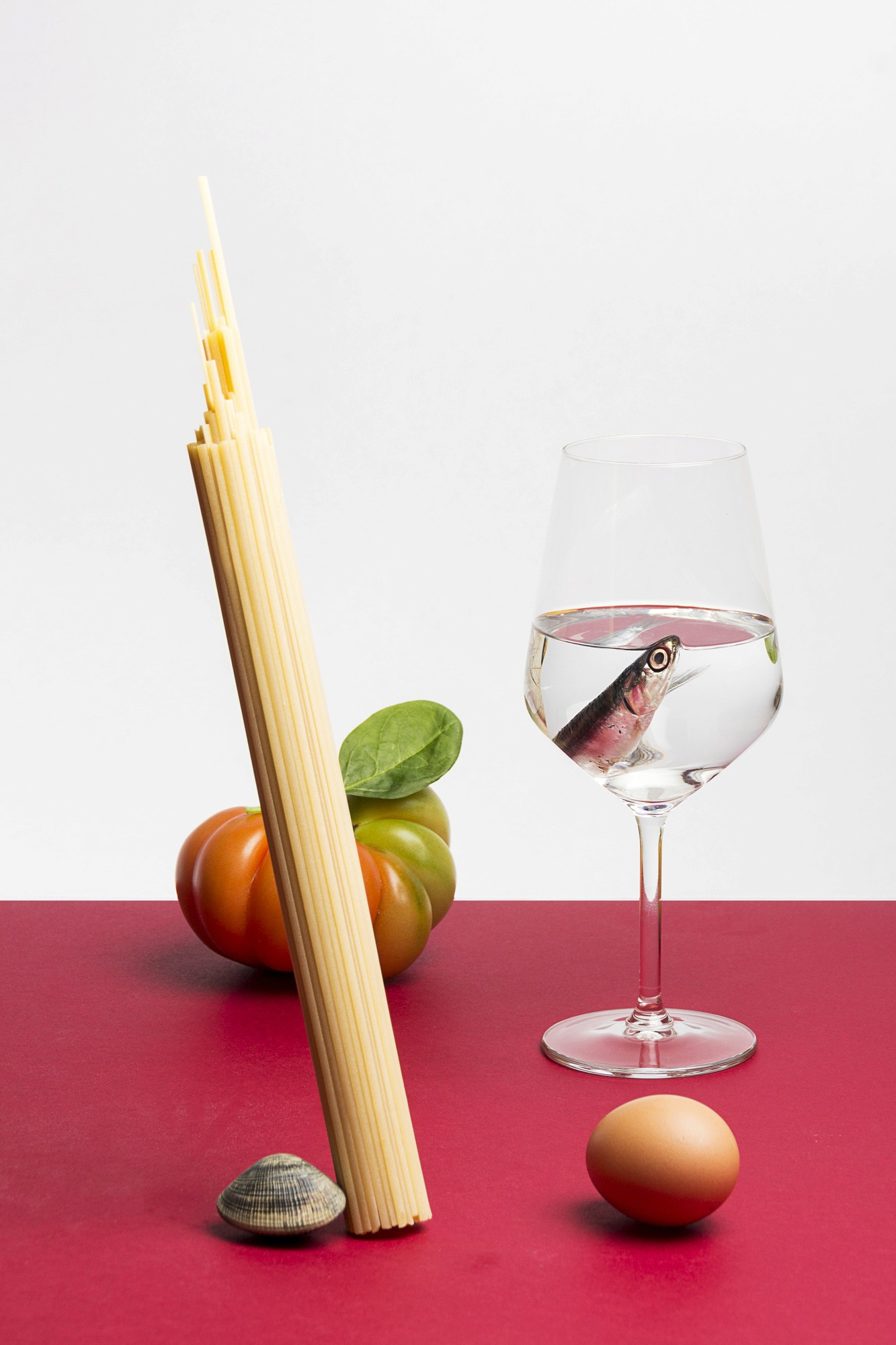 Nature morte studio sur une table au fond rouge d'ingrédients mis en scène comme des spaghettis, une tomate coeur de boeuf, un oeuf, une palourde et une sardine dans un verre à vin rempli d'eau transparente