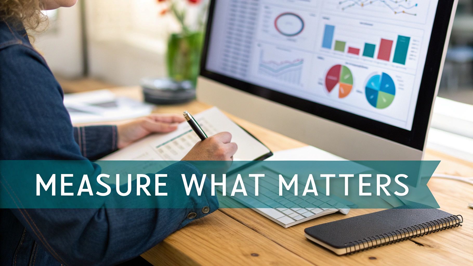 A person works at a desk, writing notes while reviewing business data on a computer monitor.