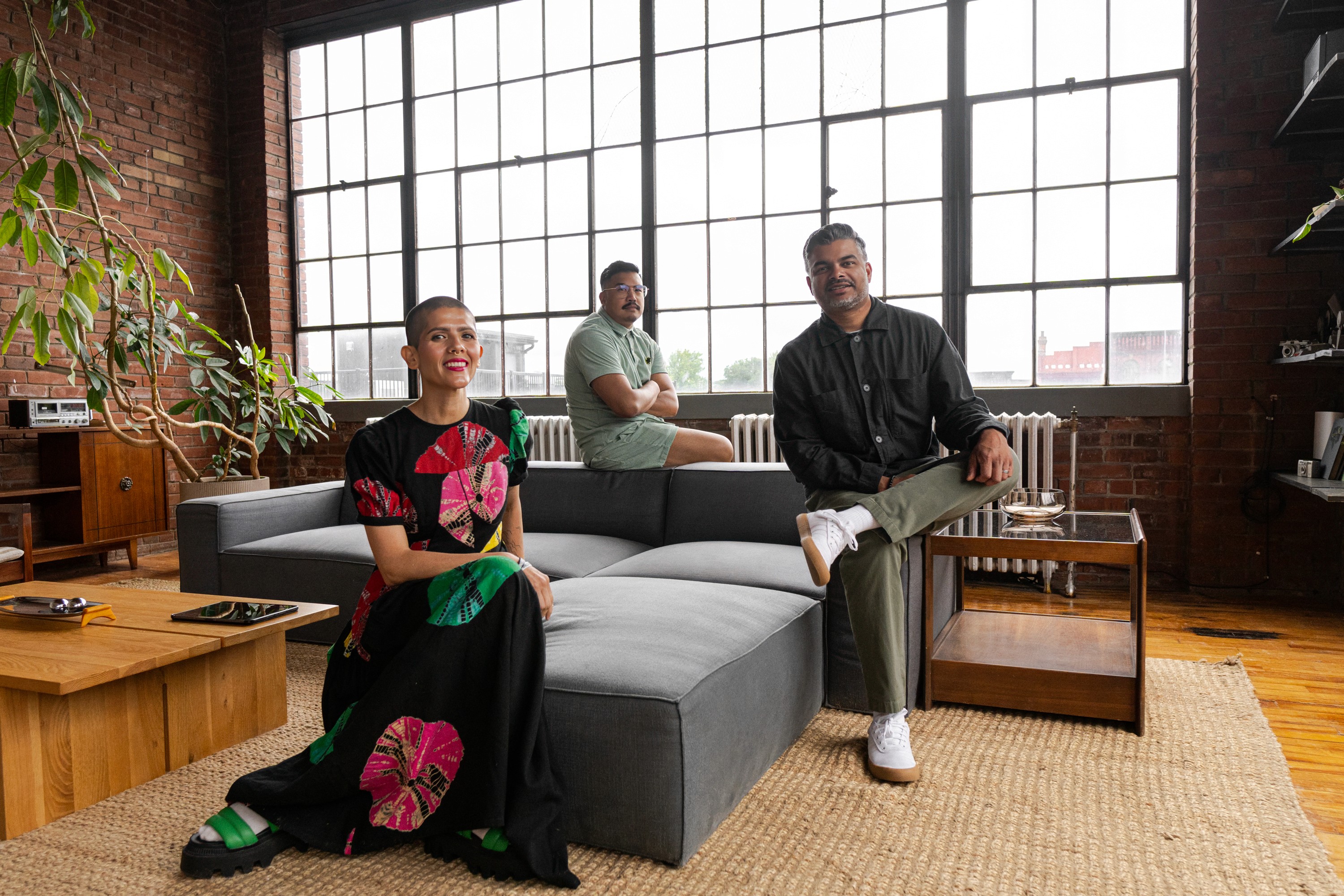 The With leadership team in a loft-style space with brick walls and large industrial windows. They are surrounded by modern furniture, plants, and natural light. Two individuals are seated on a gray sectional sofa, one wearing a green outfit and white sneakers, and the other in a colorful dress. Two others stand behind them, one wearing a gray shirt and the other in dark clothing.