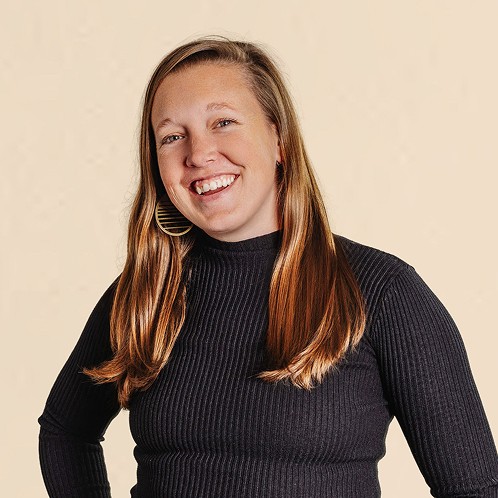 Smiling woman with long hair wearing a black ribbed top, standing against a neutral background.