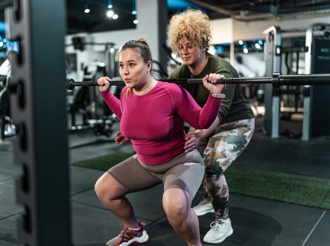 woman spotting her friend while she uses a barbell to do squats during the best gym routine for weight loss
