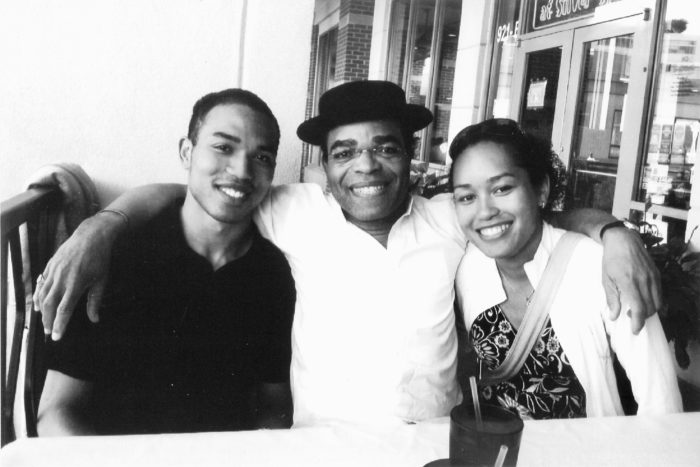 Three people smiling and sitting at an outdoor restaurant table; the man in the center wears a dark hat and has his arms around the two others in this black-and-white photo.