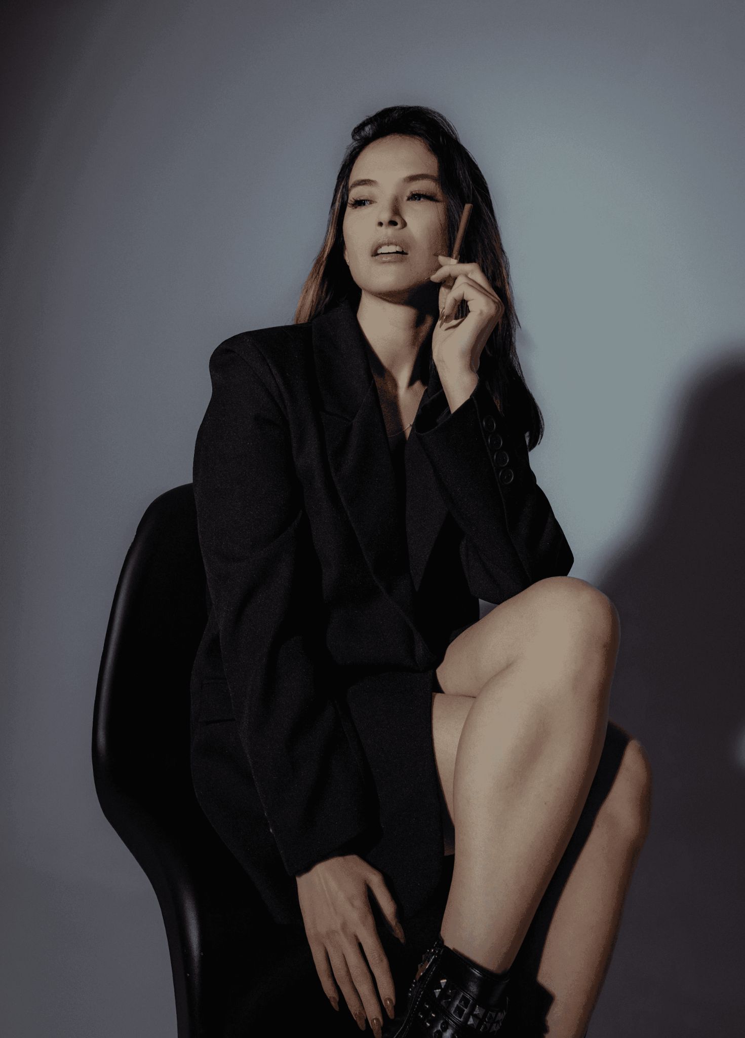 A woman in a dark, sophisticated suit sits elegantly on a black chair, holding a cigarette and gazing thoughtfully into the distance, casting a dramatic shadow against a muted background.