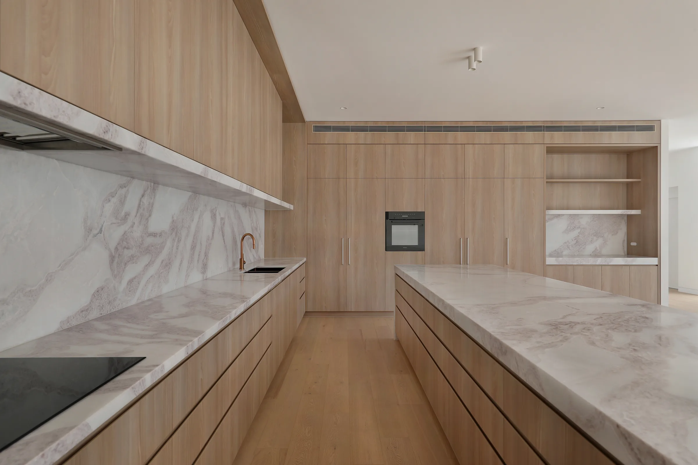 Galley kitchen with light timber cabinetry, white marble benchtops and splashback, induction cooktop, copper tap and an integrated oven