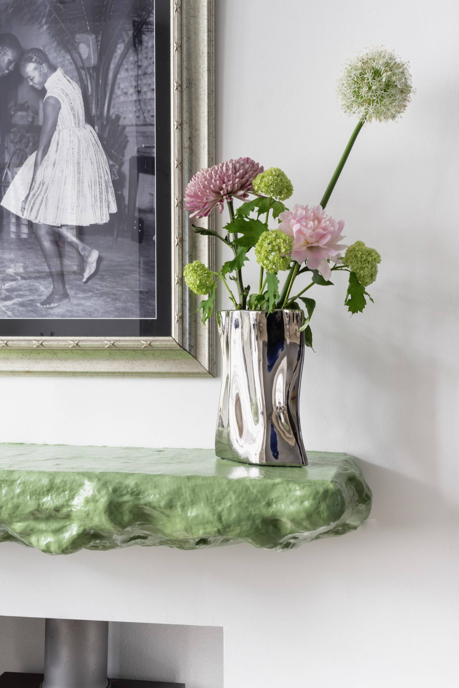 A silver, reflective, uniquely shaped vase containing pink and white flowers, placed on a green textured tabletop next to a black and white framed photograph of two women dancing.