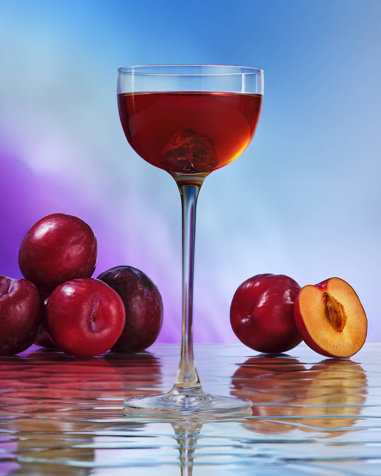 A glass of red drink stands on a reflective surface, surrounded by red apples and a slice of peach against a colorful background.