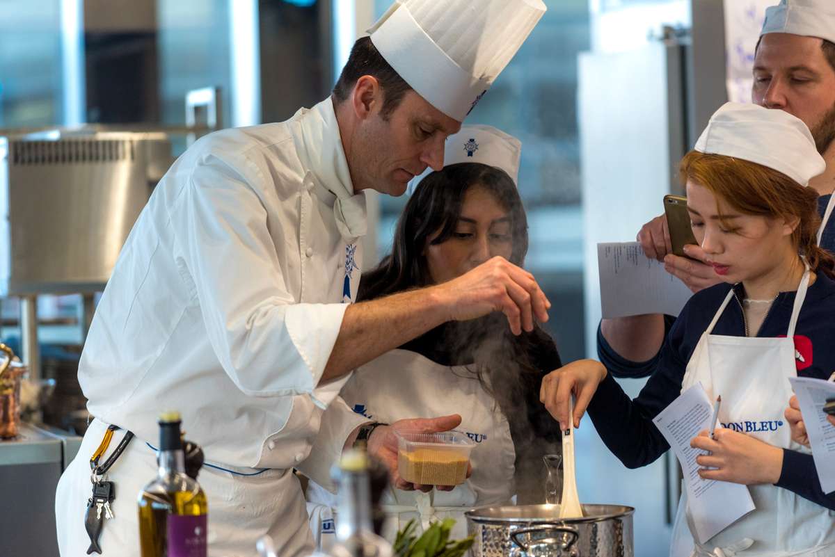 A chef and two children in white hats and aprons prepare food together in a professional kitchen.