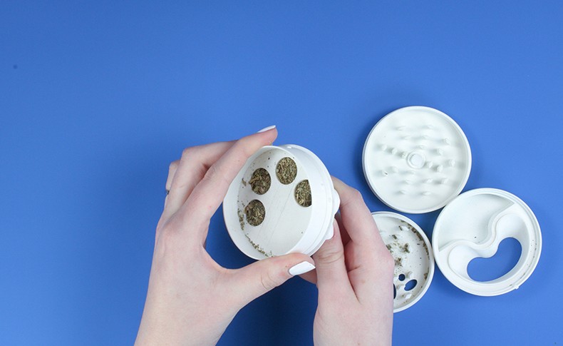 Two hands hold a white plastic container with small brown seeds inside. The container is open, revealing the seeds inside. To the right, two white plastic lid components are visible: one with a grid pattern and the other with a heart-shaped opening. The items are arranged on a blue surface.