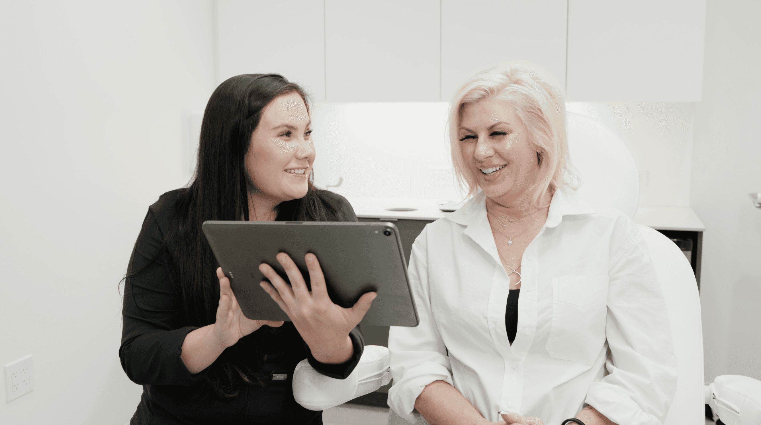 Two women are smiling and engaging with a laptop in a bright, modern workspace.