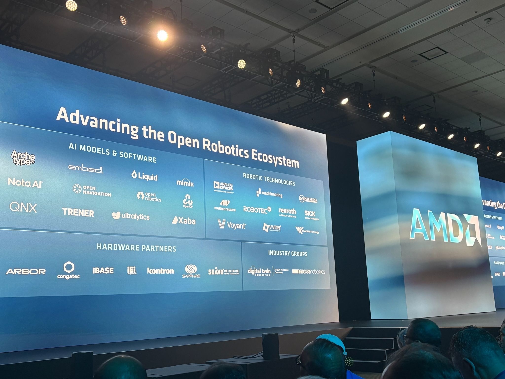 Keynote stage at AMD's Advancing AI 2026 conference, showing a slide titled "Advancing the Open Robotics Ecosystem" with Trener listed among the AI Models & Software partners, alongside Robotic Technologies, Hardware Partners, and Industry Groups categories, with the AMD logo displayed on the adjacent screen.