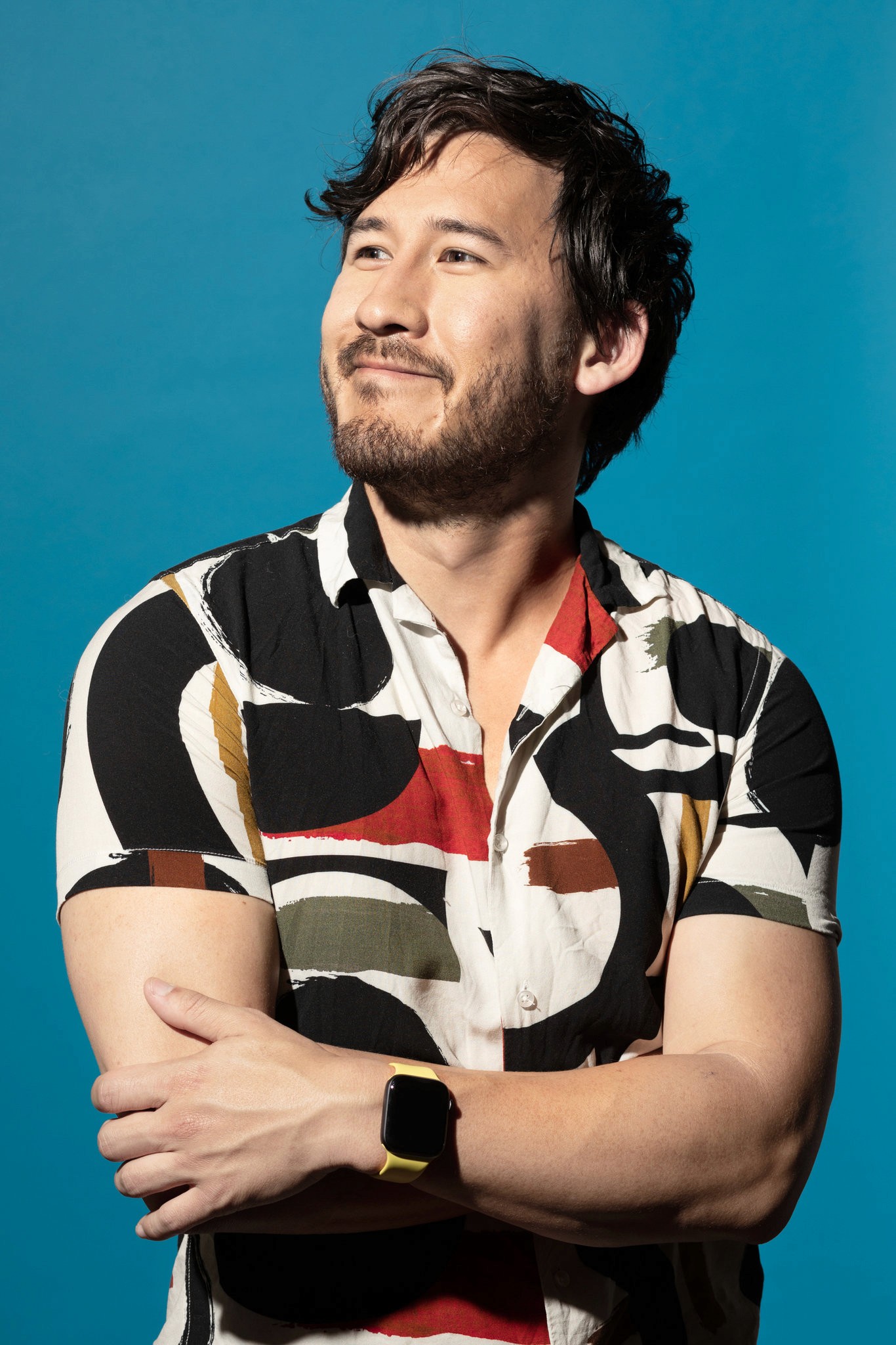 A man in a colorful patterned shirt poses confidently against a bright blue background with arms crossed.