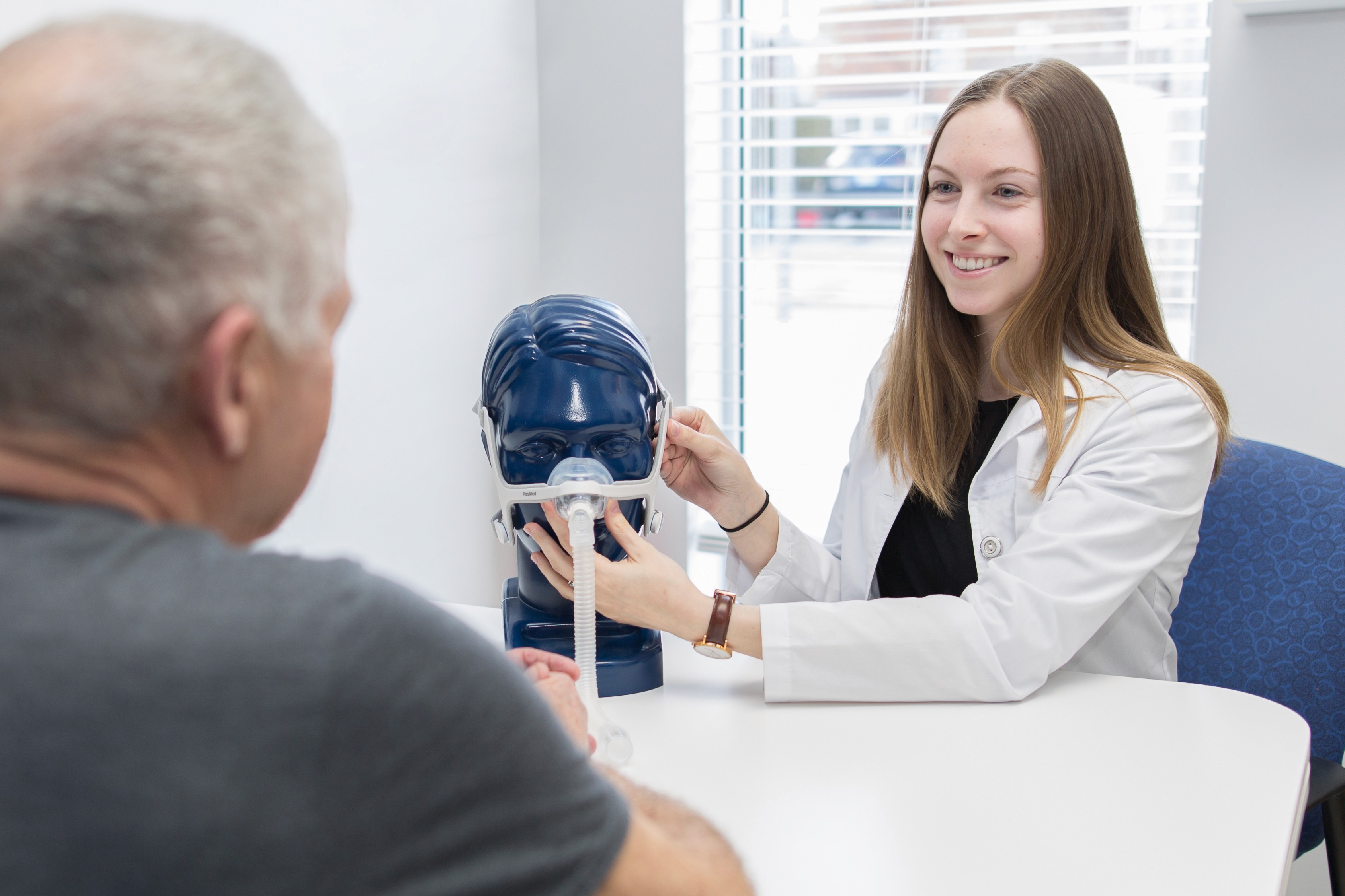 A young female medical professional explaining how a CPAP machine fits on a male bust. In the foreground a middle aged male is visible listening to her instructions. 