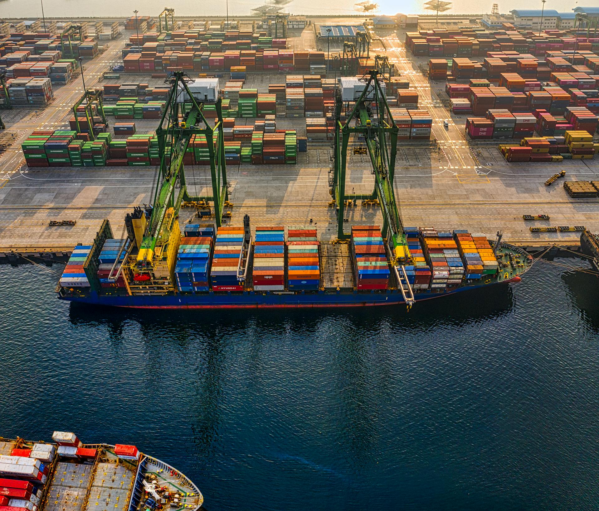 Aerial view of a container ship being loaded by cranes at a busy cargo port with stacked containers.