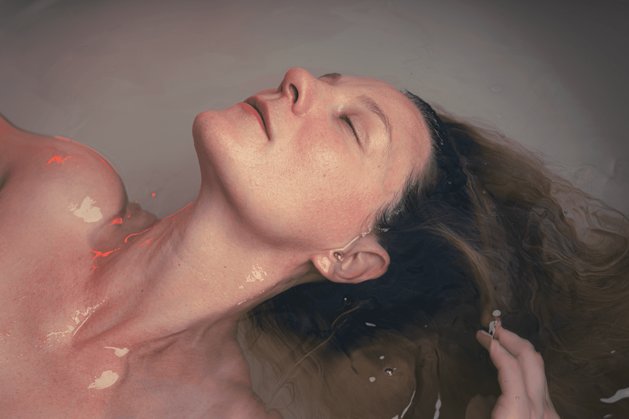 A cozy and inviting spa room with a neatly made bed, soft lighting, and warm wooden tones. The scene is partially framed by green plants, adding a natural and soothing touch.