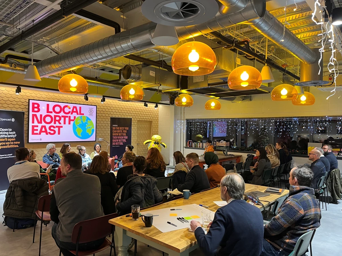 Group of people seated in a modern room with orange pendant lights, listening to a panel in front of a screen that reads “B Local North East” with an Earth graphic.