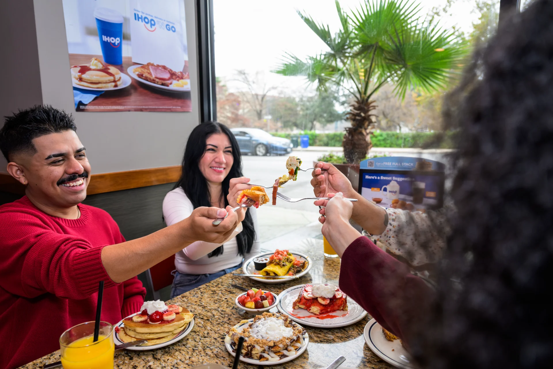 Three friends sharing pancakes, crepes, and orange juice at a window table inside IHOP