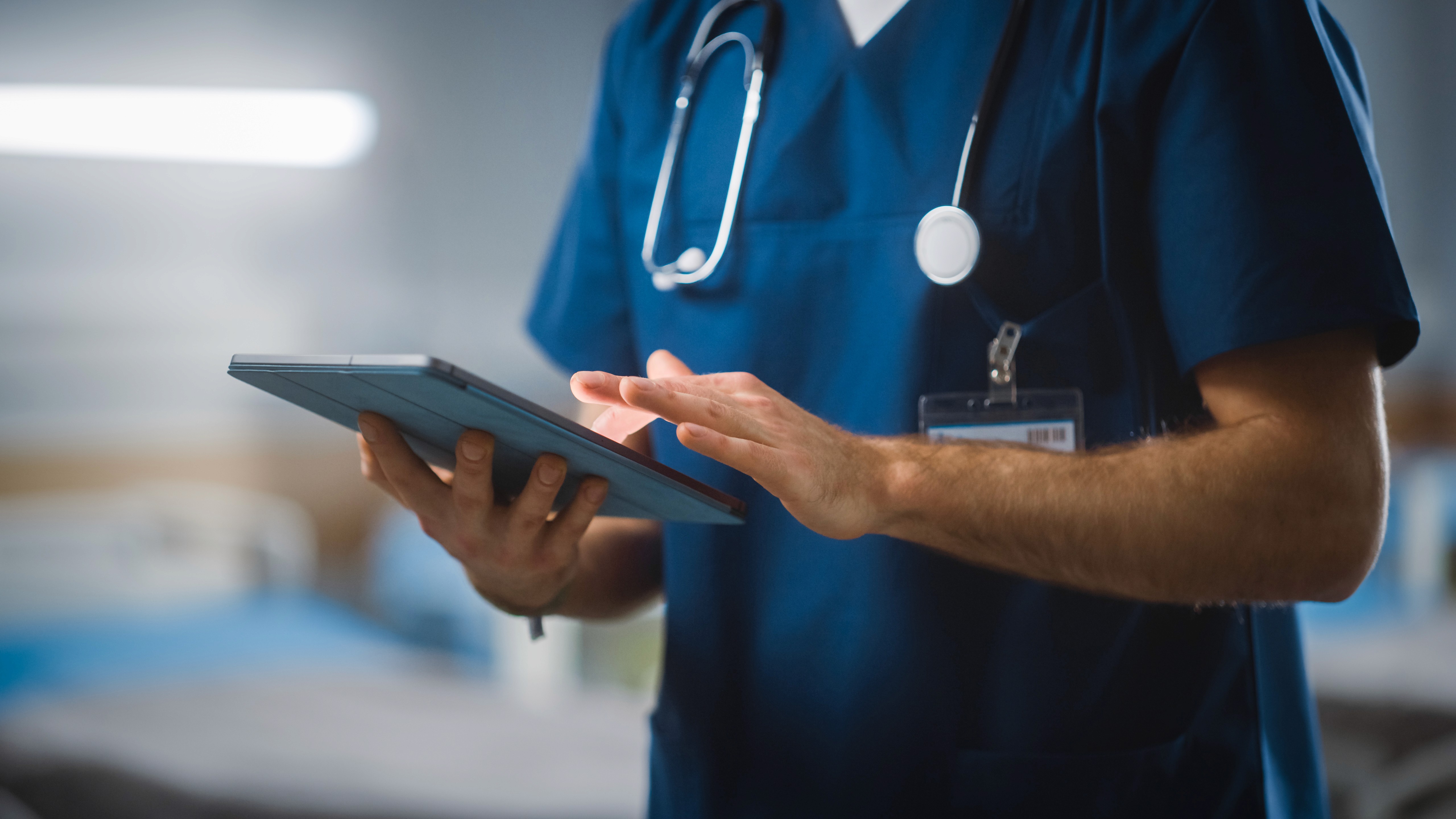 A medical professional in scrubs holding a tablet in a hospital setting, focused on the screen.