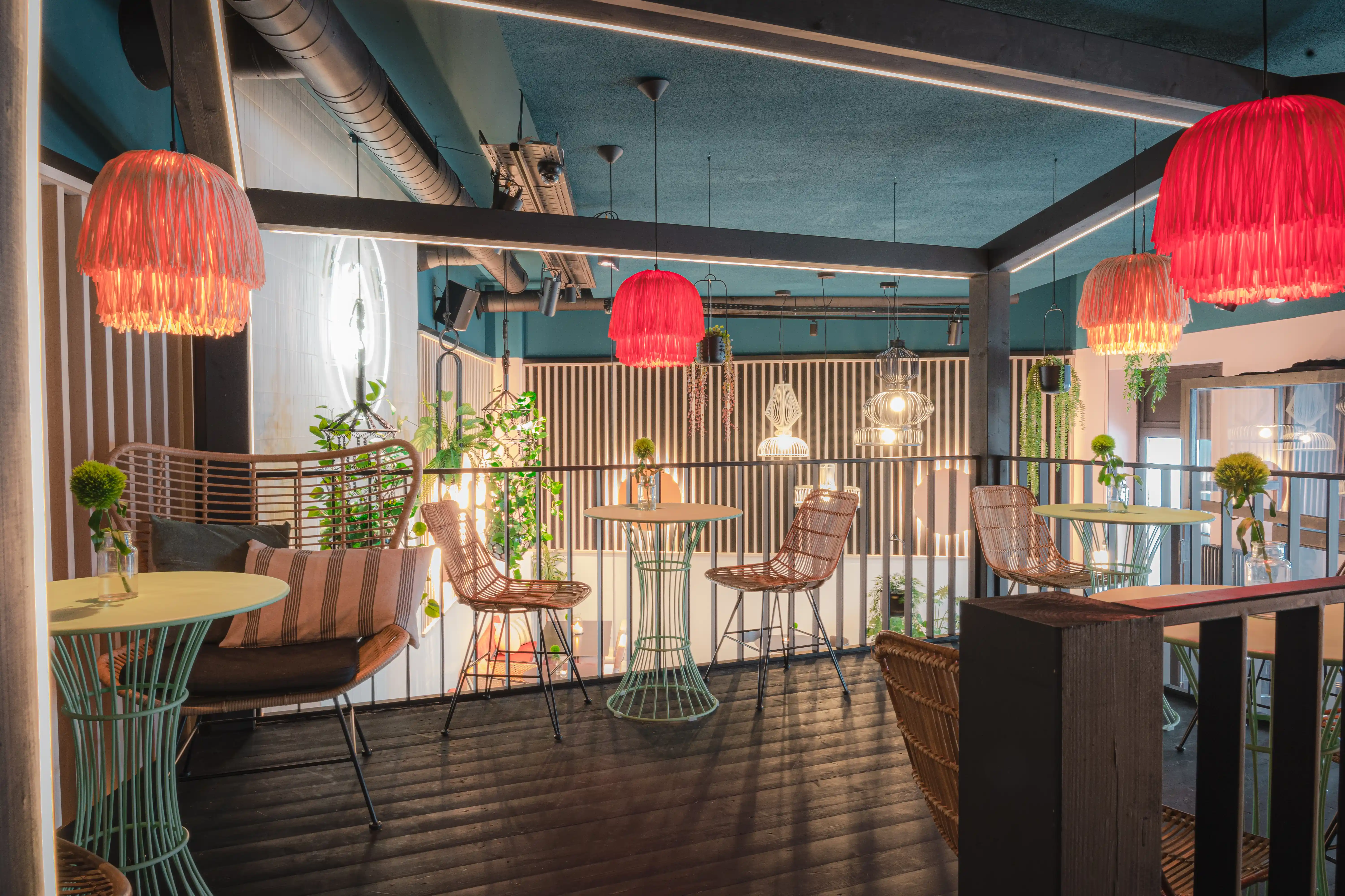 Gallery level at LOKAL VIER with rattan high chairs at small round tables, red fringe pendant lamps, striped lounge cushions and plants along the black railing.
