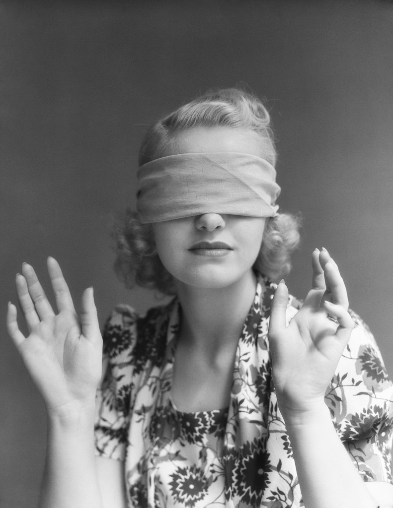 A vintage black-and-white portrait of a woman with wavy blonde hair, wearing a floral-patterned dress, blindfolded with a cloth, and raising her hands gracefully.