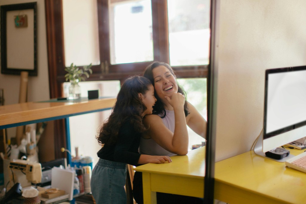 A mother and daughter share a joyful moment in a bright home office, with the daughter whispering in the mother's ear as they laugh together near a yellow desk and computer.