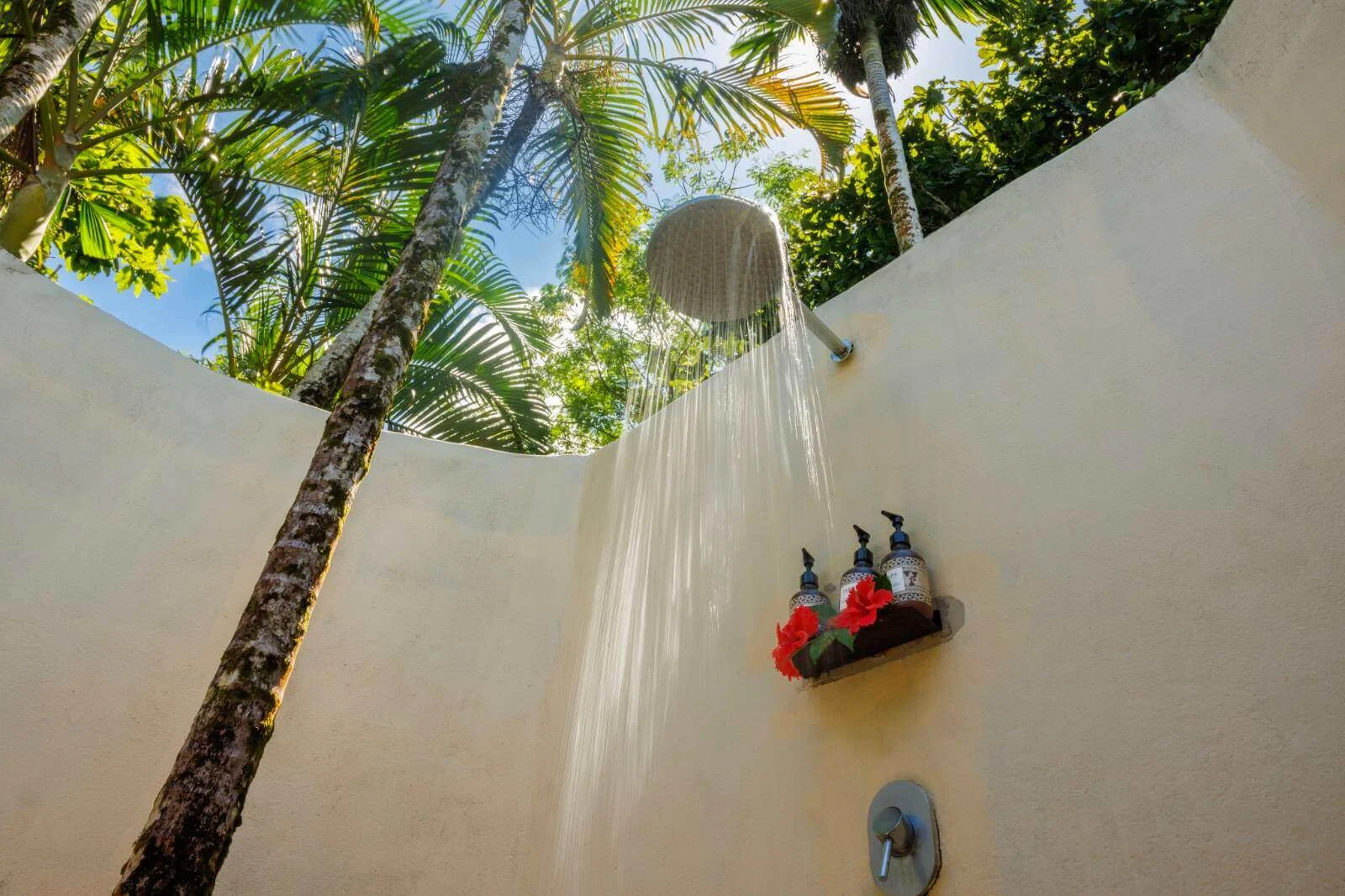 Outdoor rain shower flowing water at Uprising Beach Resort, surrounded by palm trees and decorated with red hibiscus.