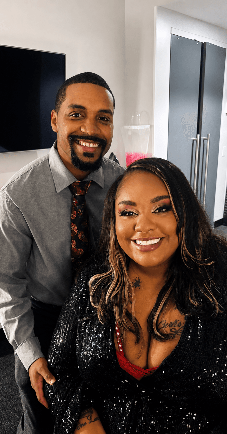 Smiling couple dressed formally indoors, posing together; woman in black sequined outfit, man in gray shirt and floral tie.