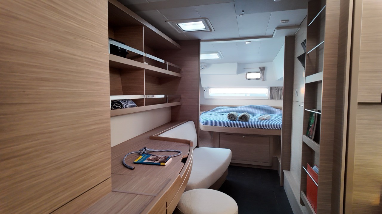 Interior view of a Lagoon 42 guest cabin, showing a berth made up with blue-striped linens and rolled towels, wood shelving, a built-in desk with cushioned bench, and a hull window.