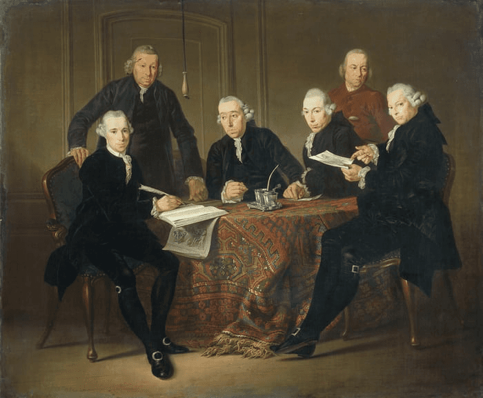 A group of six men in historical clothing gather around a table, engaged in conversation and discussion.
