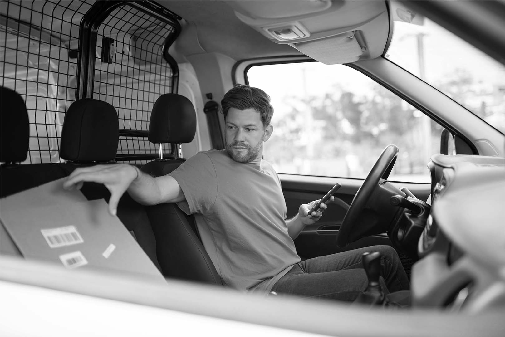 A man sitting in the driver’s seat of a van, looking over at a box package on the passenger seat. The setting suggests a delivery or transportation context