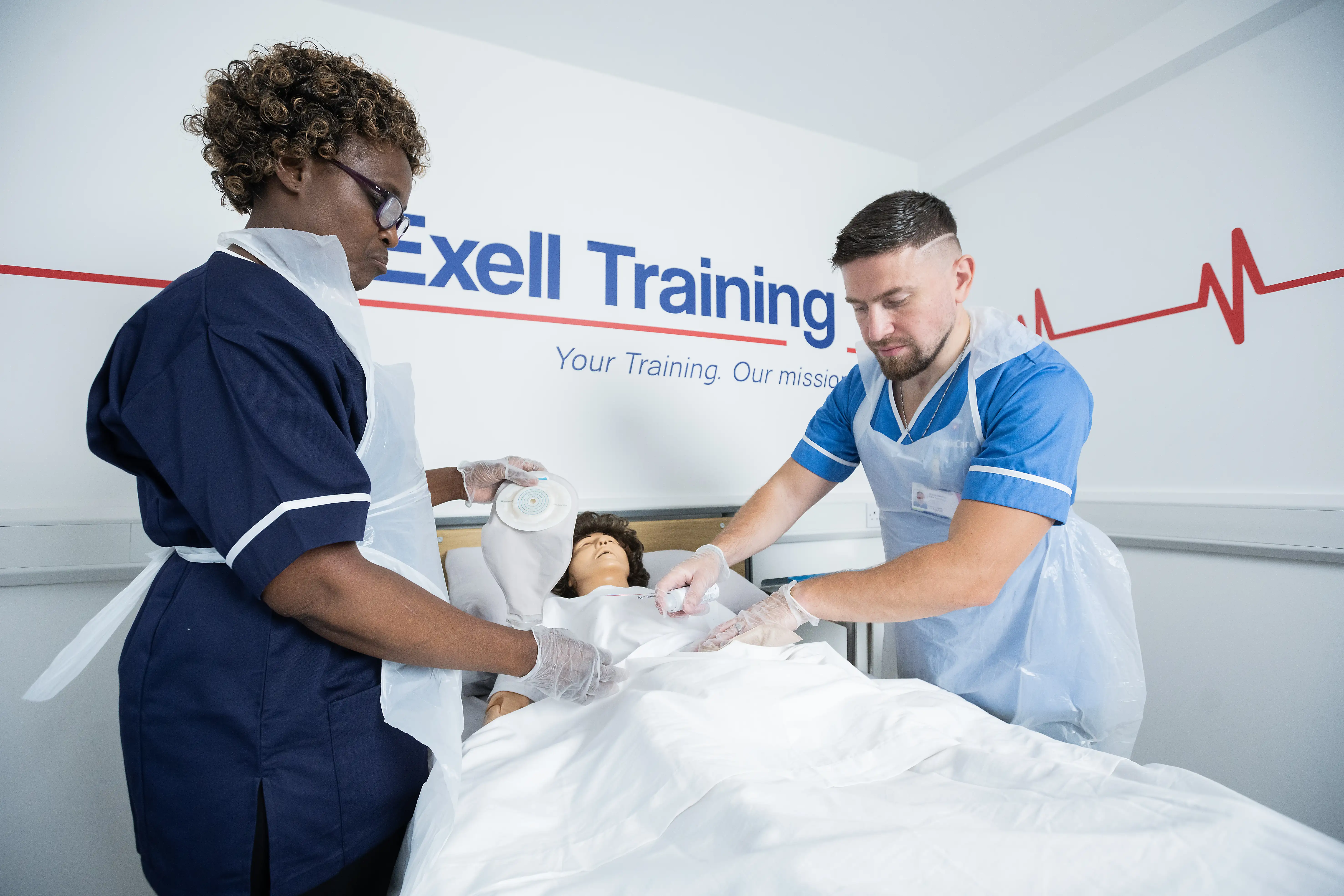 Two healthcare workers practise stoma care on a mannequin during a training session at Exell Training. They are wearing gloves and aprons while applying and positioning stoma equipment.