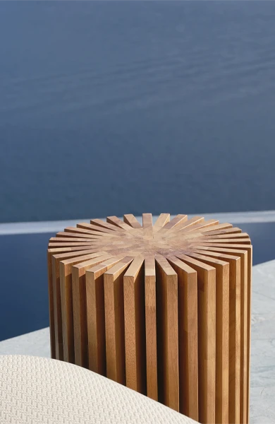 Round outdoor side table in solid Iroko wood, crafted with interlocking vertical panels in a radial pattern to form a sculptural cylindrical shape – featured poolside in a luxury modern setting with lounge chairs.
