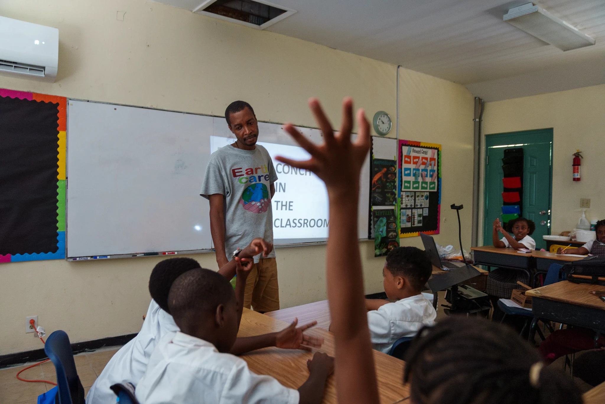 A teacher stands at the front of a classroom near a whiteboard, engaging with students. One student raises their hand while others are seated around desks. Posters and a clock are visible on the walls.