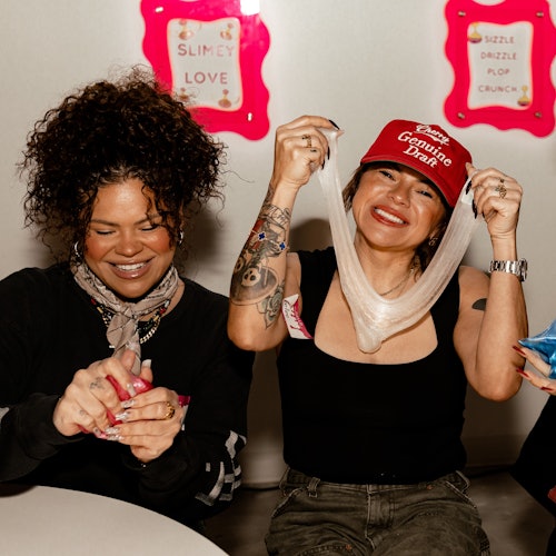 Dos mujeres sonriendo y jugando con slime; una lleva una gorra roja y la otra lleva una bufanda. Carteles brillantes con texto al fondo.