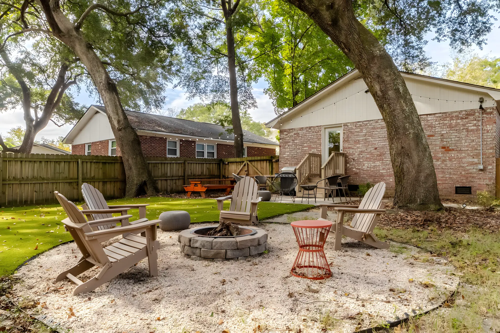 Stone fire pit ringed by Adirondack chairs under two big oaks