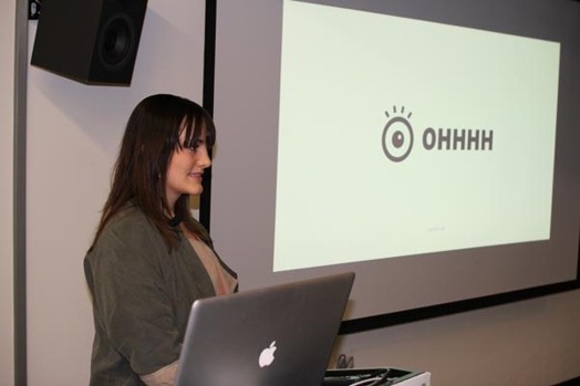 A photo of a woman giving a lecture from a laptop while on the screen projector there is an eye pictogram and the word OHHH.