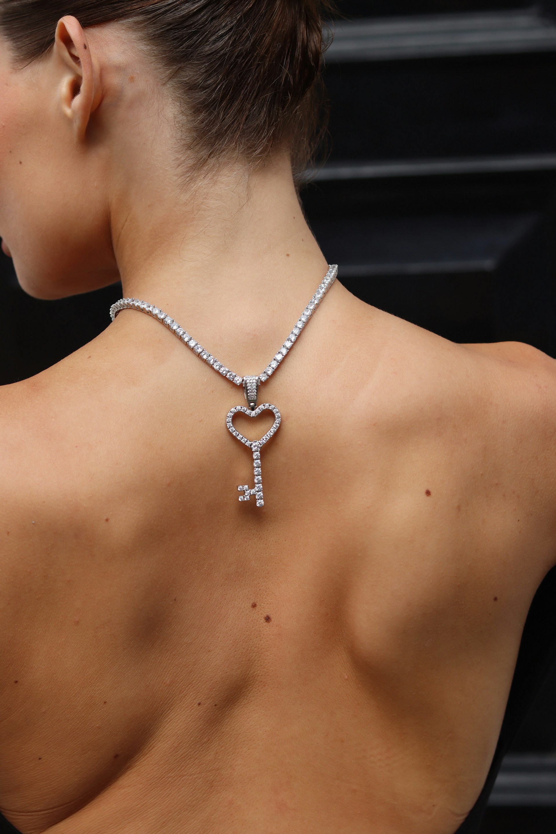A silver-toned tennis chain with a large diamond-encrusted heart-shaped key pendant draped over a woman's bare back.