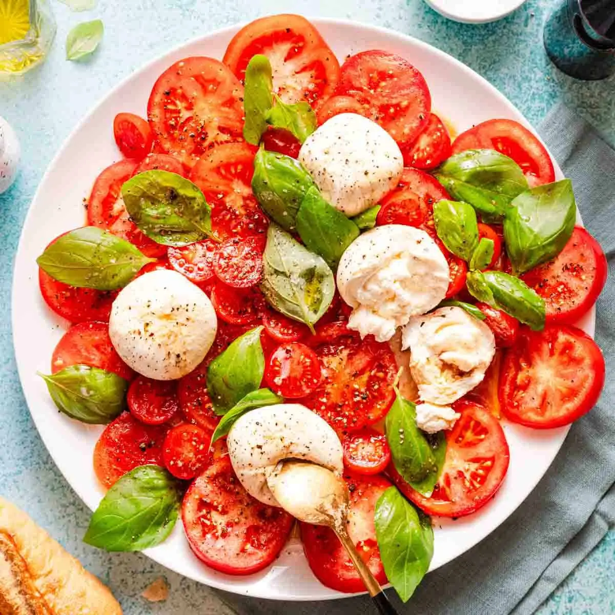 Plate of burrata caprese salad topped with whole and torn burrata, sliced tomatoes, cherry tomatoes, fresh basil leaves, olive oil, and black pepper.
