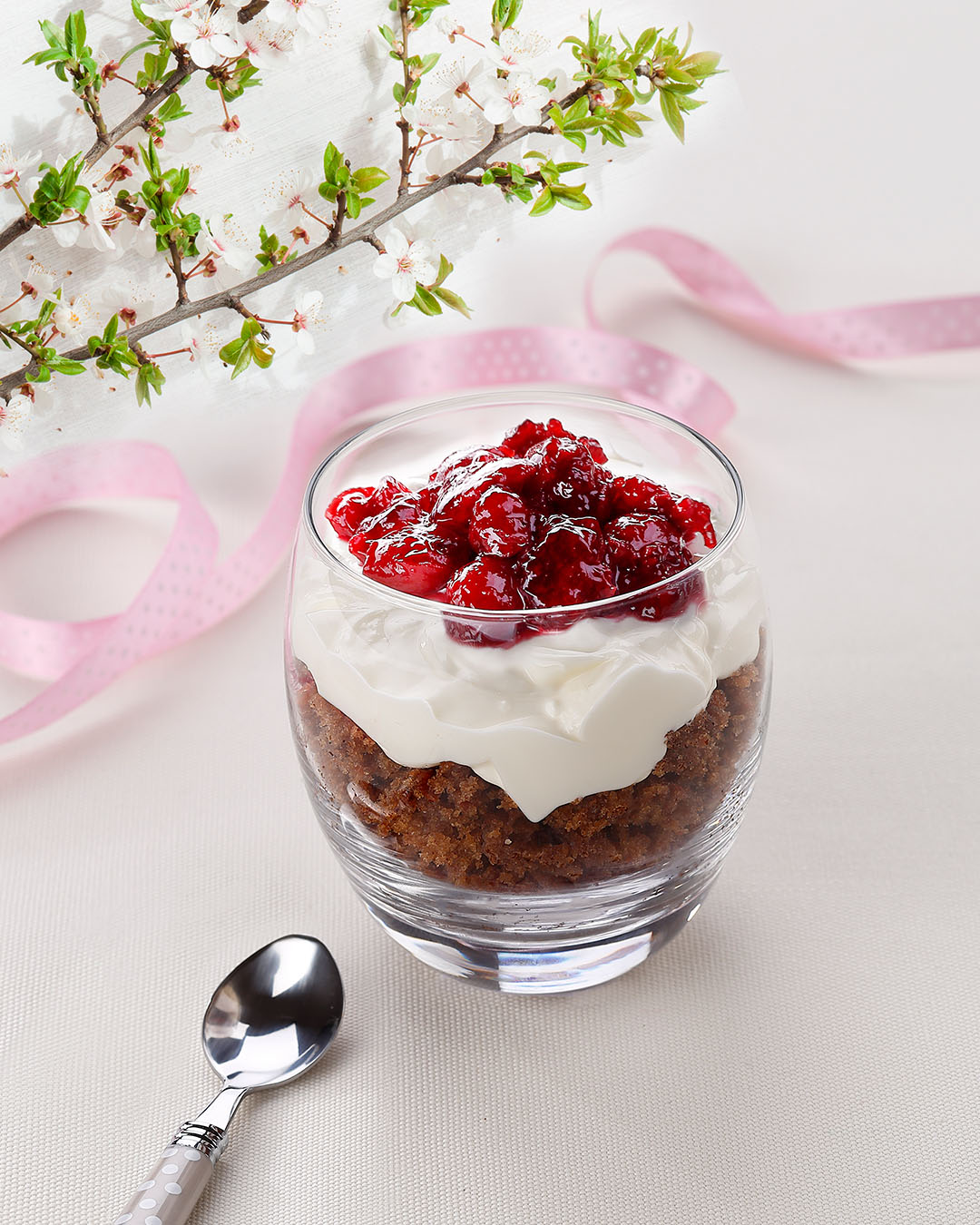 A glass dessert bowl filled with layers of rich chocolate cake, whipped cream and a red cherry garnish sits on a light tablecloth, accompanied by a silver spoon and decorated with a pink ribbon and blooming white flowers to give the dish a spring feel.