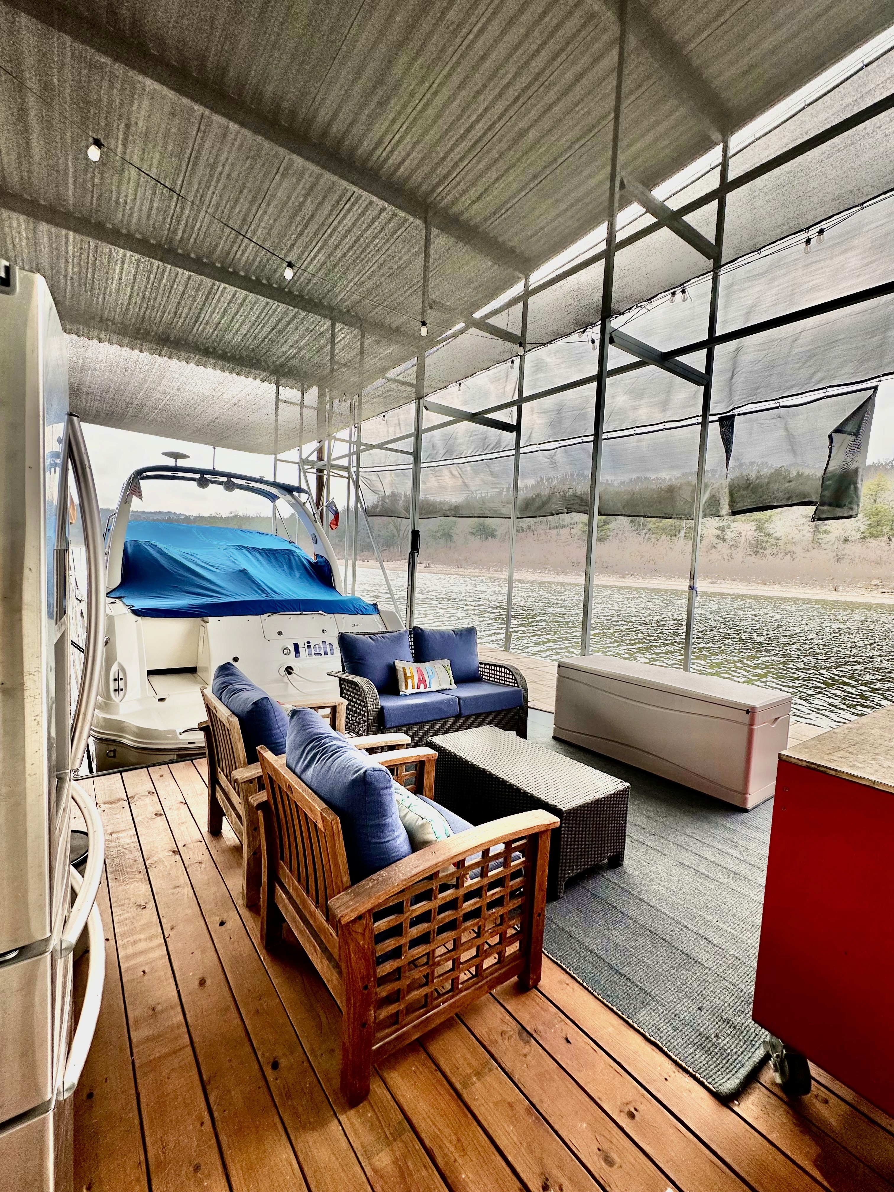 A covered dock with a wooden deck features cozy outdoor seating, including cushioned armchairs and a sofa, positioned next to a boat with a blue cover, providing a comfortable waterside retreat.