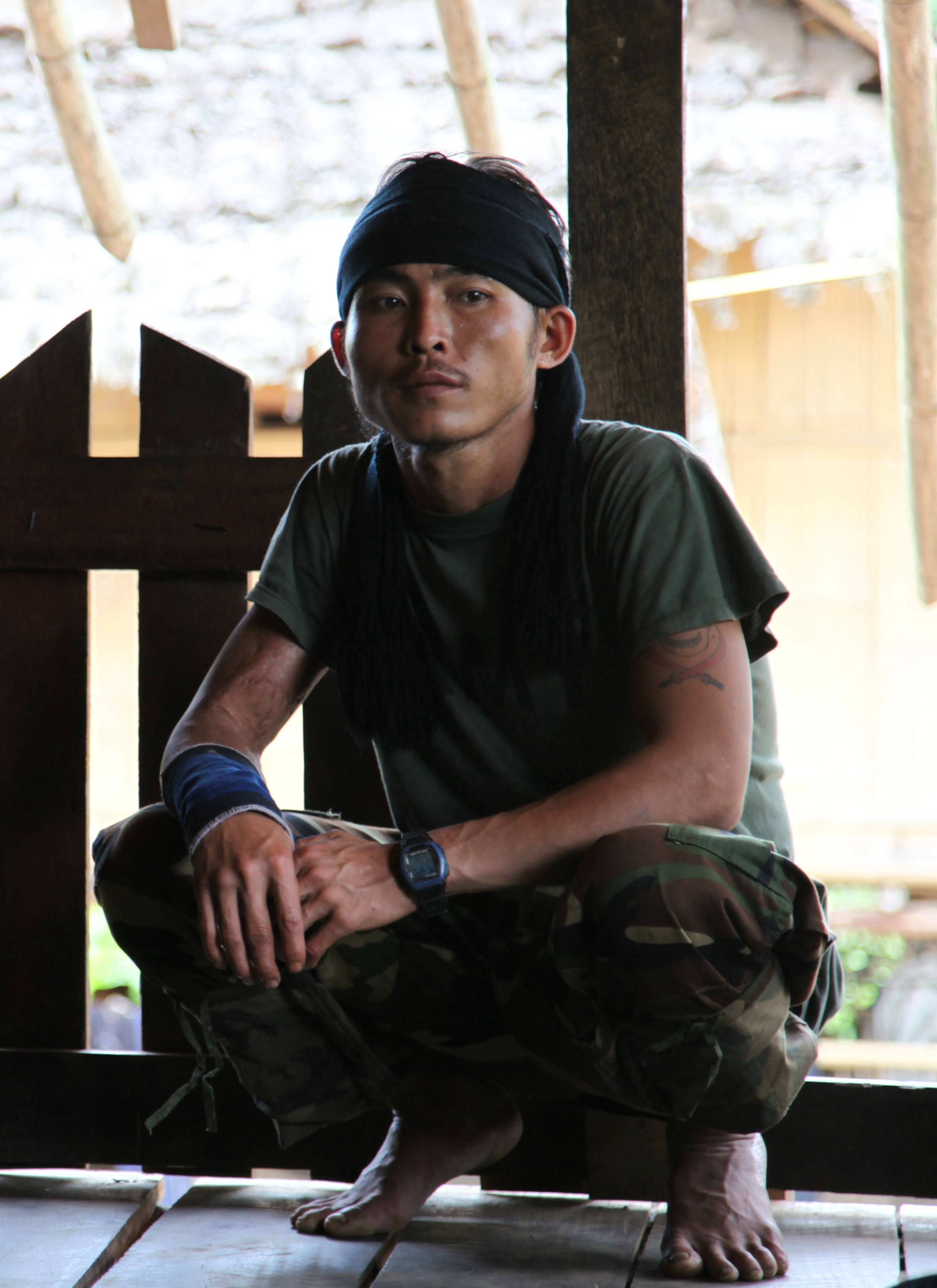 A man squats barefoot on a wooden floor inside a rustic structure. He wears camouflage pants, a dark green T-shirt, and a black headband with long fabric trailing down. A blue wrist wrap and black digital watch are visible on his left arm. Natural light filters in from behind, illuminating parts of his face and surroundings.