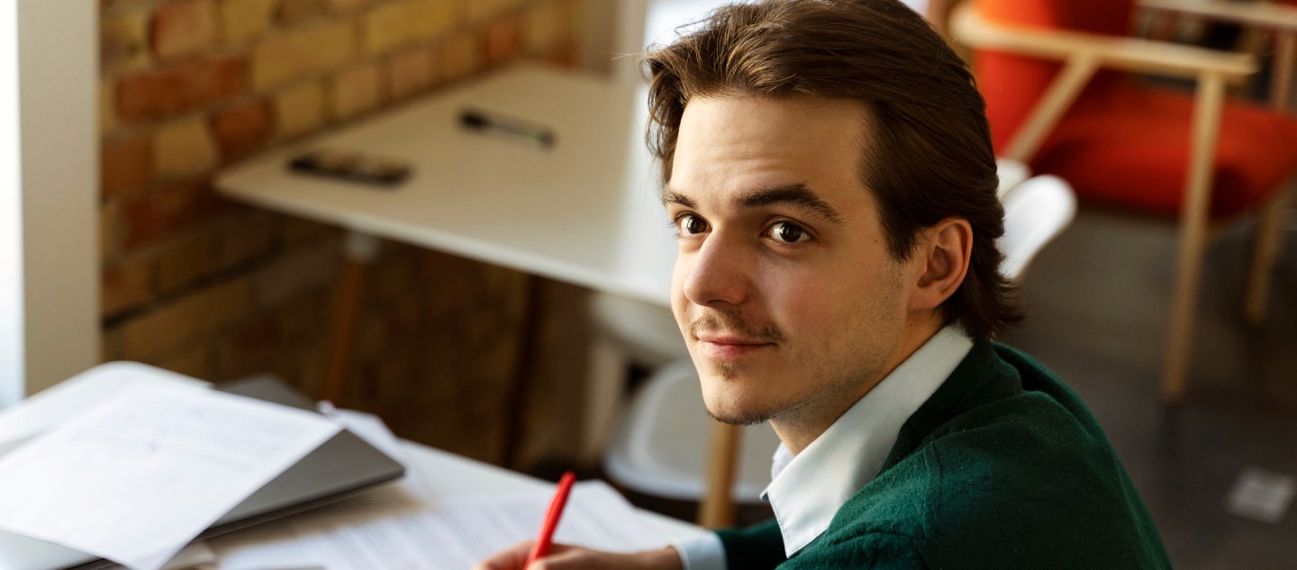 A person with short brown hair wearing a green sweater sits at a desk with scattered papers and a red pen in hand, against a backdrop of a brick wall and modern furniture. 